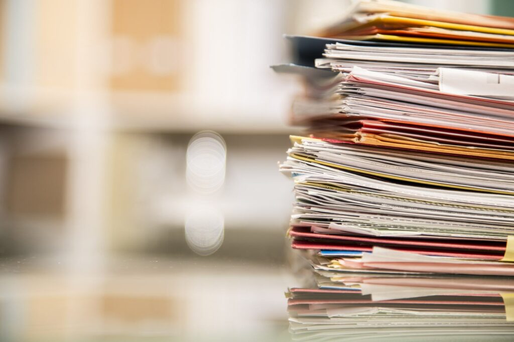 large stack of file folders, documents, paperwork piled on glass top desk in office