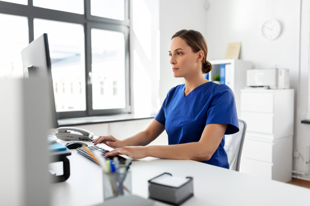 nurse sitting at a desk in a hospital in front of a desktop computer wearing blue scrubs and the room around her is monochromatic and white