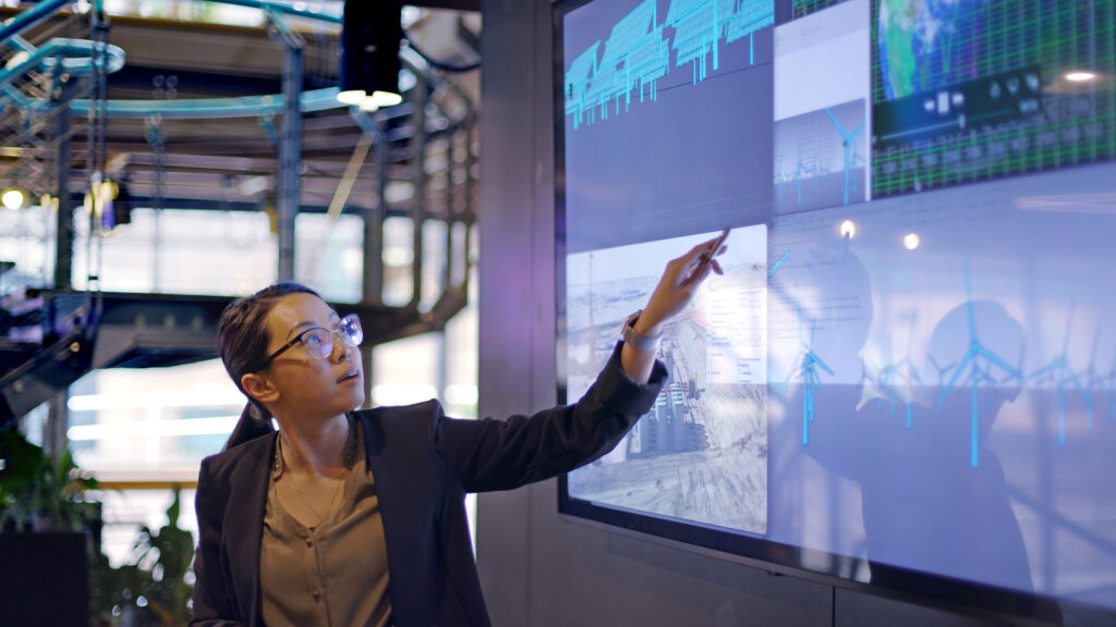 young woman conducting a lecture with the aid of a large screen displaying data and designs concerning low carbon electricity production with solar panels and wind turbines